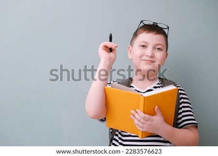 A cute cheerful boy in stripped tee shirt is holding a yellow book and a black pen and school bag on back on green background.