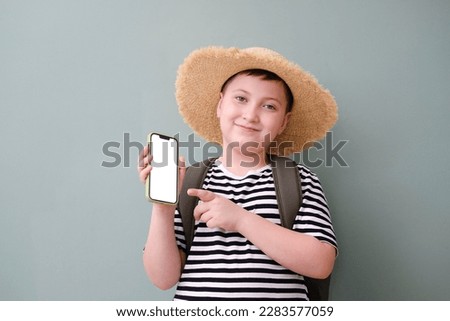 A cute cheerful boy in stripped tee shirt and in straw hat with bag holding and showing by thumb on the white screen of mobile phone on green background.