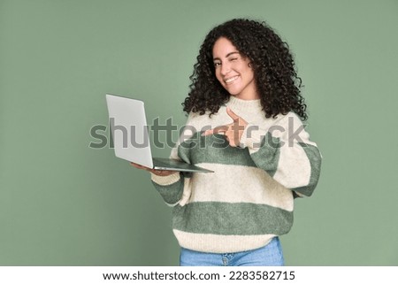 Young happy latin woman winking pointing at laptop isolated on green background. Smiling female model holding computer presenting advertising new trendy website job search or dating service.