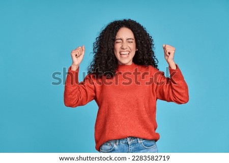 Young happy latin woman winner screaming celebrating crazy money win isolated on blue background. Excited female model feeling euphoric lucky about win or achievement with yes gesture.