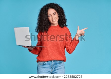 Young happy latin woman holding laptop pointing aside isolated on blue background. Smiling female model holding computer presenting advertising job search or ecommerce shopping website.