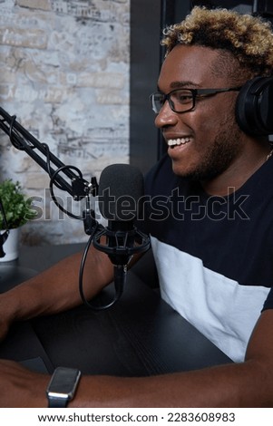 Young guy listens to an online lecture, uses a laptop and headphones to communicate with the lecturer and classmates. Joyful black radio presenter in a modern loft-style studio hosts the program. 
