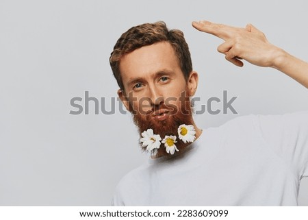 Portrait of a funny man in a white T-shirt with flowers daisies in his beard on a white isolated background, copy place. Holiday concept and congratulations.