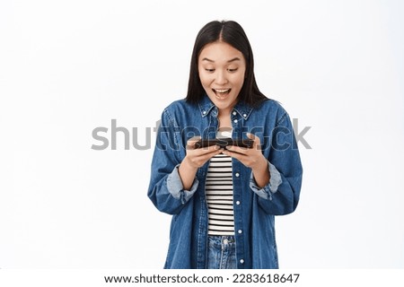Image of asian girl watches video on smartphone, looks at mobile phone amazed, playing video game, stands over white background. Royalty-Free Stock Photo #2283618647