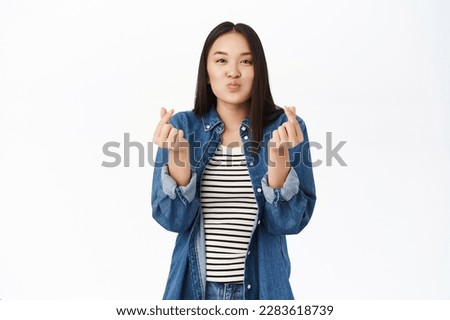 Cute young woman, 20 years old, asian girl shows heart finger sign, I love you gestures, stands over white background.