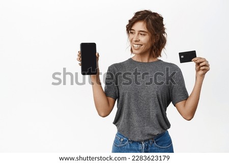 Portrait of cute girl with phone, looks and shows smartphone screen, holds black credit card, white background.