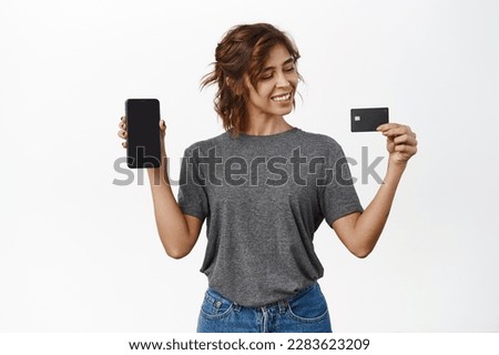Smiling young woman shows mobile phone screen, looks satisfied at credit card, standing in grey t-shirt over white background.