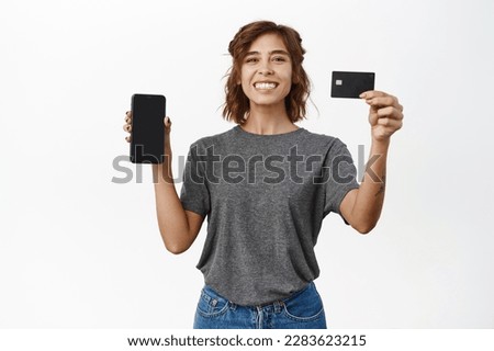 Smiling young woman showing mobile phone screen, app interface, recommending credit card, standing in t-shirt over white background.