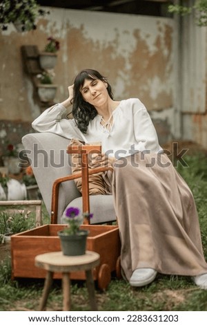 a young attractive brunette woman with long hair is sitting in a cozy armchair in the courtyard of a country house