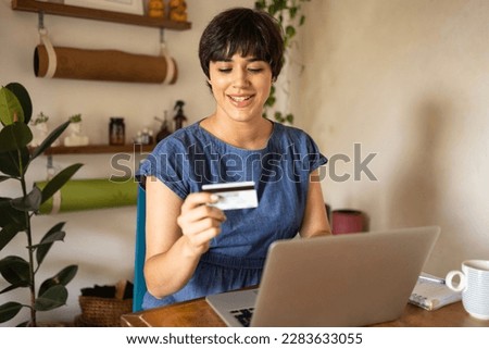 Young latin girl with short hair smiling while holding a credit card. She is sitting in apartment full of green plants and using a laptop. Royalty-Free Stock Photo #2283633055