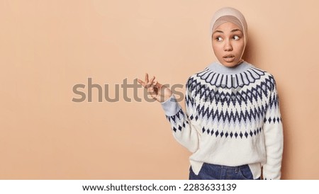 Confused hesitant Muslim woman has confused expression raises hand and being doubtful wears hijab and jumper isolated over brown background with copy space for your advertisement. Doubt concept