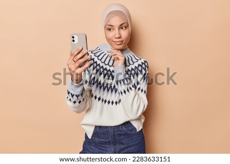 Calm beautiful Muslim woman with gentle expression keeps hand under chin has healthy skin focused at front camera of smartphone for making selfie wears traditional veil warm jumper and jeans