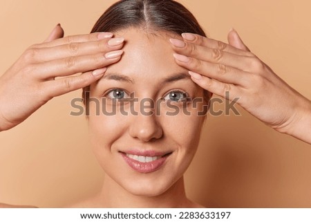 Close up shot of dark haired young woman examines facial skin keeps hands on forehead looks directly at camera has natural beauty manicure isolated over brown background. Photo taken in studio