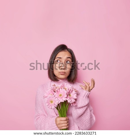 Vertical shot of dark haired Asian woman feels confused looks unaware above holds bouquet of gerbera flowers wears knitted jumper poses against pink background blank space your promotional text