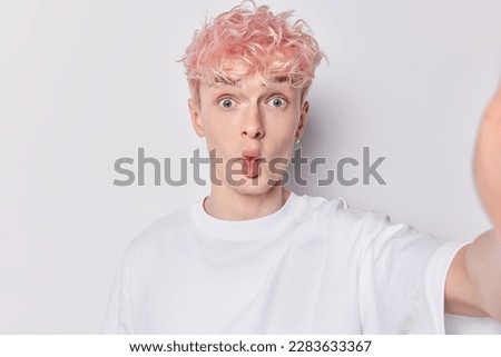 Surprised guy with pink hair puckers lips shows fish mouth as foolishes around keeps arm outstretched pose sfor selfie dressed in casual t shirt isolated over white background. Facial expressions Royalty-Free Stock Photo #2283633367