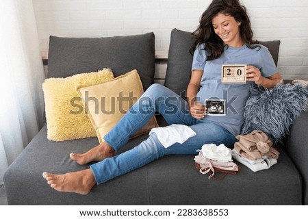 Beautiful pregnant woman looking a cute baby clothes and ultrasound picture at home. Anticipation of a child birth.