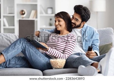 Relaxed happy cheerful young multicultural spouses chilling together on couch at home at weekend, looking at laptop screen and smiling, eating popcorn, arab guy holding remote control