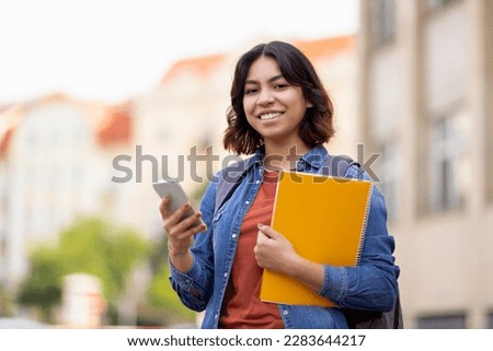 Outdoor Portrait Of Smiling Arab Female Student With Smartphone And Workbooks, Happy Young Middle Eastern Female Using Mobile Phone While Standing Outside At Campus, Resting After Classes