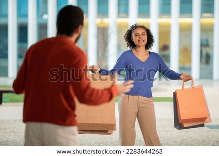 Unrecognizable Husband Reacting To Shopaholic Wife Posing With Paper Shopping Bags And Smiling Standing Outdoors. Selective Focus On Female Buyer. Shopaholism Issue Concept