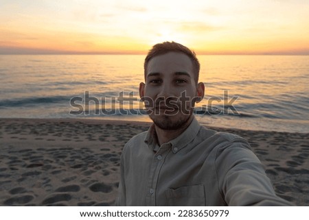 young man in a shirt, taking a selfie in a sunset on the beach