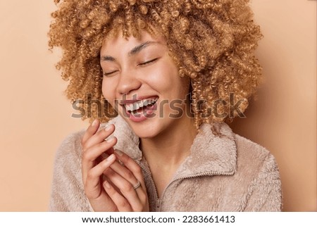 Close up shot of positive curly haired woman laughs happily keeps hands together closes eyes from pleasure dressed in winter jacket remembers something funny from life isolated over brown background Royalty-Free Stock Photo #2283661413