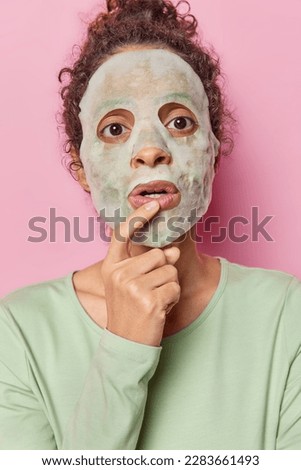 Face care concept. Surprised curly haired woman applies beauty sheet mask for cleaning skin keeps finger on lips wears casual green jumper poses over pink background undergoes cosmetology procedures