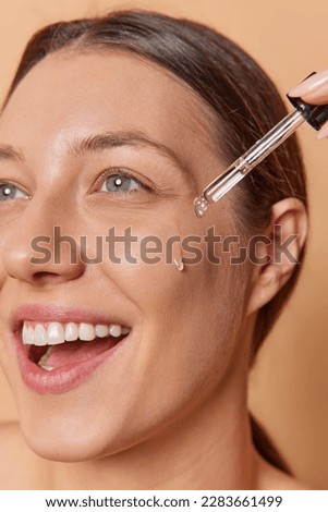 Vertical shot of happy young woman applies moisturizing face serum or essential oil with pipette enjoys new cosmetic product and self care routine smiles toothily poses indoor. Skin care concept