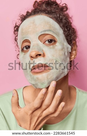 Headshot of serious woman applies moisturising sheet mask does skin care procedures touches face looks attentively at camera isolated over pink background background. Beauty procedures concept