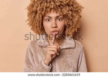 Serious strict woman with curly hair makes silence gesture demands to keep information in secret has scared surprised expression wears jacket isolated over brown background. Dont drop any word