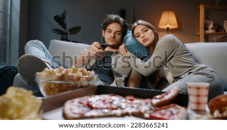 Young caucasian couple extremely frustrated. Bored students are sitting in front of tv, switching channels with remote and eating unhealthy food, tired after hard day 
