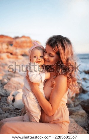 Young mother with her daughter on vacation in the summer on the beach. Mom kissing the baby. Rocky beach, mindful motherhood, baby care, mother's day, first year of a baby's life, parent. Family look
