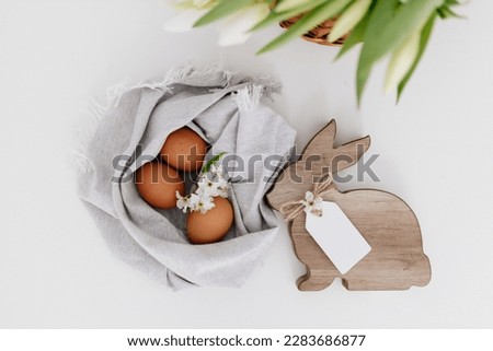 A wooden Easter bunny with a signature tag sits near brown eggs, which lie in a natural linen napkin and white tulips on a white background.  Easter card. Spring background