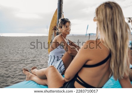 Beautiful girls in a swimsuit with a sports body sitting under the wooden canopy on a sand beach. Two tanned young woman resting and sunbathing on a holiday on the sea shore. Copy space.