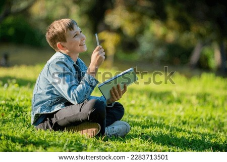 Smiling school boy with a textbook in his hands is sitting on a green lawn in a park. Pupil studies in the park while sunny day. A thinking teen boy with  notebook in a garden looking up. Royalty-Free Stock Photo #2283713501