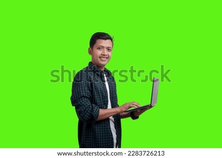 Happy and smiling millennial Asian man in plaid suit holding laptop looking at camera while standing on green studio background