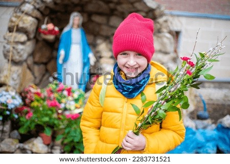 a happy boy in warm clothes holds a bouquet with willow and flowers, stands in front of a figure or sculpture, a statue of the virgin mary on palm Sunday