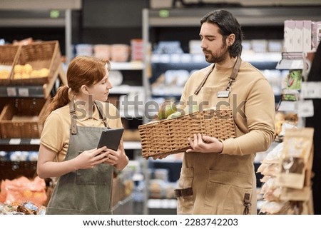 Waist up portrait of two young workers in supermarket stocking shelves with fresh fruit
