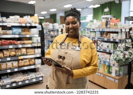 Waist up portrait of smiling black woman working in supermarket and looking at camera cheerfully