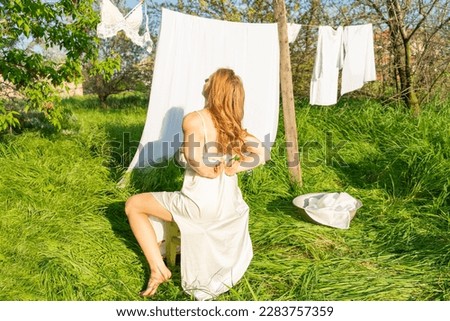 beautiful red girl in nightie hanging laundry outdoors. village woman working in countryside.Cute girl in dress washing white clothes in metal basin in backyard, hanging laundry on clothesline
