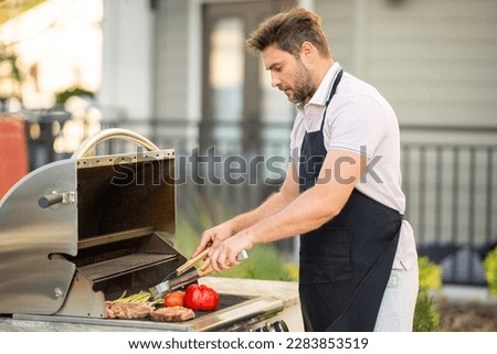 Man cooking meat on barbecue in the backyard of the house. Handsome man preparing barbecue. Barbecue chef master. Man in apron preparing delicious grilled barbecue food, bbq meat. Grill and barbeque. Royalty-Free Stock Photo #2283853519