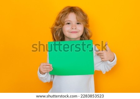 Kid showing index finger on green empty sheet of paper, isolated on yellow background. Portrait of a kid holding a blank placard, poster.