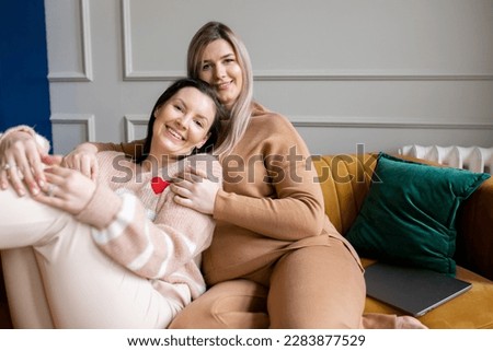 Happy women in textile suits sit on stylish mustard color sofa hugging each other in living room. Friends have fun and relax on female meeting. Home leisure, lifestyle, only women.
