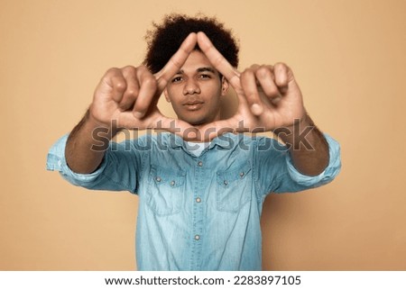 Portrait of handsome young man with dark skin and afro hair in blue denim shirt making and showing triangle gesture on brown background. Environment protection, recycling and reusing concept Royalty-Free Stock Photo #2283897105