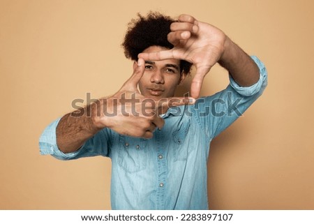 Young latin dark-skinned guy showing camera shape gesture with hands looking through at camera, picking best position for making picture, isolated on brown studio background in denim clothes