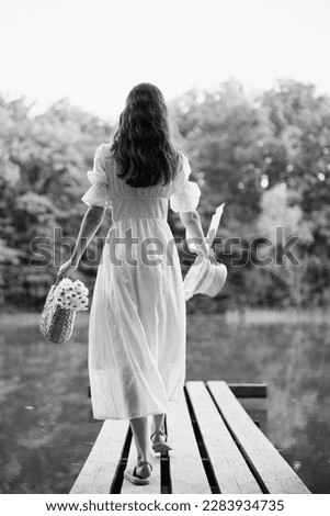 a woman in a long light dress stands on a pier by a lake in the forest with a basket and a wicker hat. monochrome photography