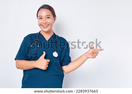 Smiling young Asian female professional nurse working wearing a blue uniform pointing a finger at empty copy space and showing thumbs up sign isolated on white background. Healthcare medicine concept