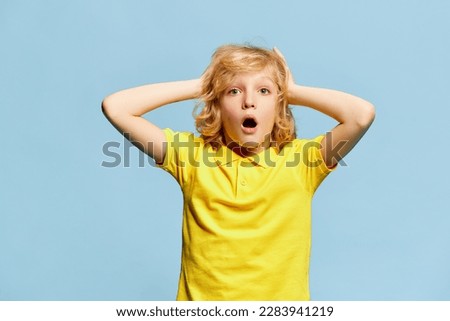 Feeling surprised. Little boy, child with blonde curly hair and shocked face posing against blue studio background. Concept of childhood, emotions, facial expression, lifestyle. Ad