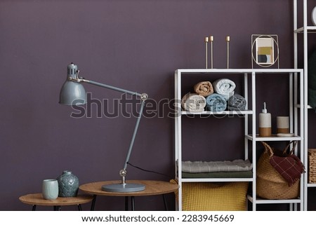 Lamp and two small vases standing on round wooden tables by wall against rack of shelves with rolled towels, picture frames and basket
