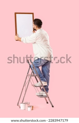 Young man with ladder hanging frame on pink background