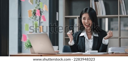 Happy young creative Asian woman working office desk feeling excitement raising her arms up as victory sign, celebrates career ladder promotion or reward. Great results successful work concept
